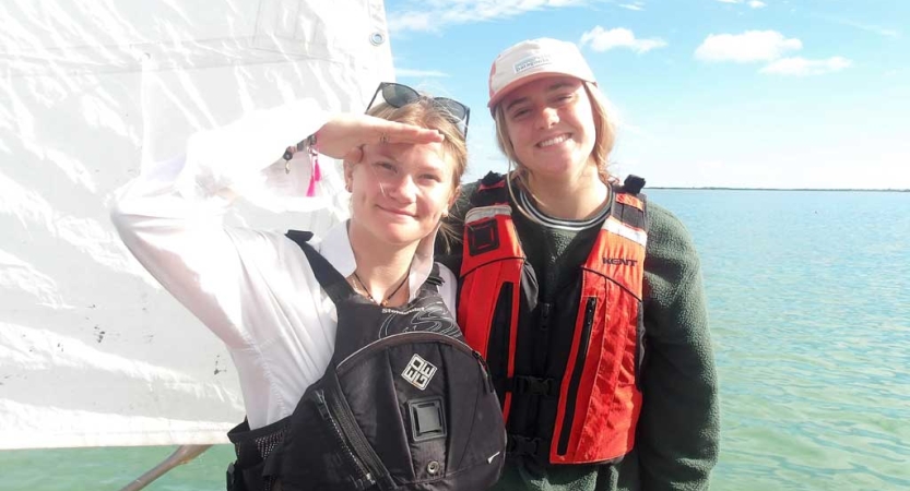 Two people wearing life jackets smile while standing on a sailboat on calm blue water. One of them is saluting the camera. 